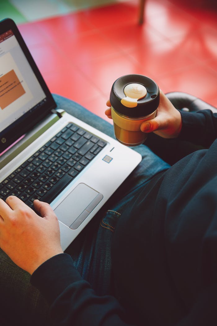 A person types on a laptop while holding a reusable coffee cup, illustrating modern remote work.